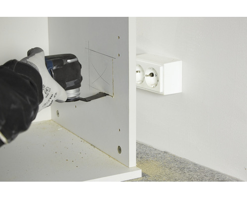 Iemand zaagt met een multifunctioneel gereedschap een gat in een houten plank naast een stopcontact.