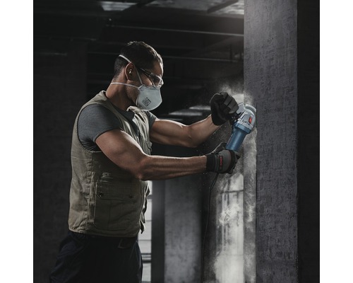 Een man met een beschermend masker en veiligheidsbril werkt met een haakse slijper op een betonnen muur.