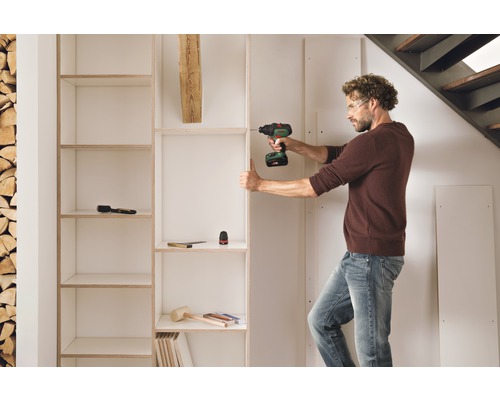 Een man monteert een witte boekenkast met een accuschroevendraaier in een kamer met een stapel hout en een trap.