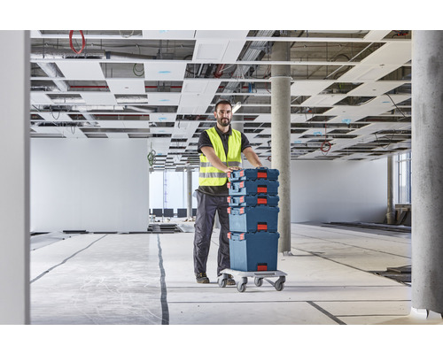 Ambachtsman met gereedschapskoffers op een rolbord in een casco gebouw.