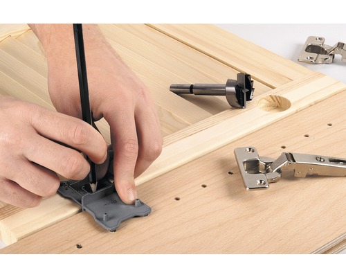 Iemand markeert de positie voor een scharnier met een sjabloon en een potlood op een houten plank, naast een forstnerboor en een scharnier.