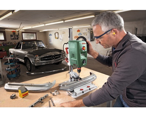 Werkplaatsopstelling met een Bosch tafelboormachine die een werkstuk boort, een man draagt een veiligheidsbril en gehoorbescherming.