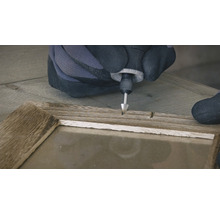 Een persoon met handschoenen gebruikt een multifunctioneel gereedschap op een houten frame.
