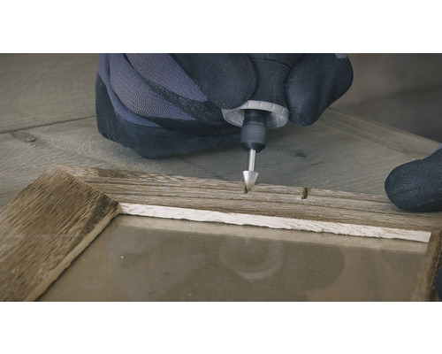 Een persoon met handschoenen gebruikt een multifunctioneel gereedschap op een houten frame.