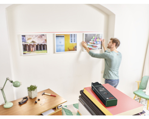Een man hangt met behulp van een laser afstands meter schilderijen aan een muur. Gereedschap ligt op een tafel.