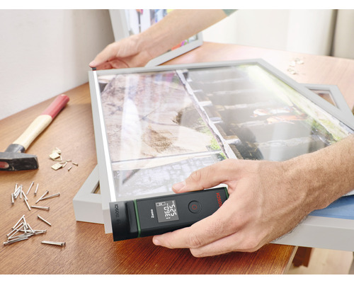 Iemand meet een fotolijst met een digitale laser afstands meter. Hamer en spijkers liggen op tafel.