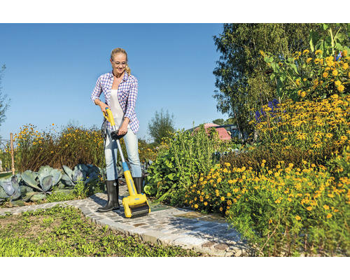 Vrouw verwijdert onkruid met een elektrische voegenborstel op een tuinpad.