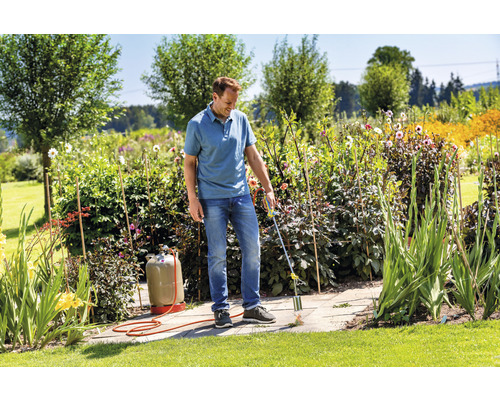 Een man verwijdert onkruid met een gasbrander op een stenen terras in een tuin.