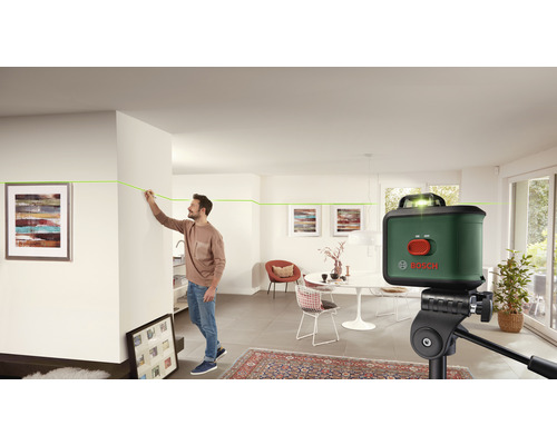 Kamerfoto met een man die met een kruislijnlaser lijnen op een muur uitlijnt, op de voorgrond een statief met een kruislijnlaser en op de achtergrond een woonkamer.