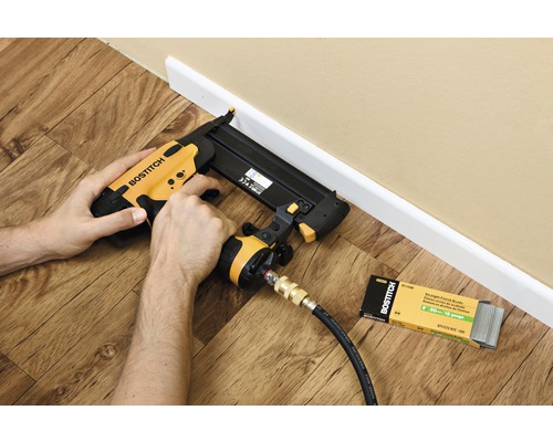 Iemand gebruikt een pneumatische tacker om een plint te bevestigen. Er ligt een pakje bijbehorende spijkers naast.