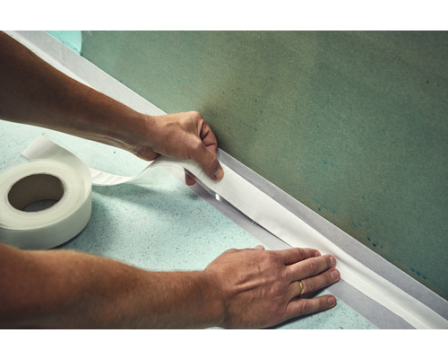 Een persoon brengt afdichtband aan op een muur. Op de vloer ligt een rol met afdichtband.