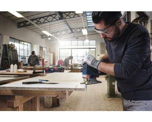 Een man gebruikt een decoupeerzaag om een houten plank te zagen in een werkplaats, met een veiligheidsbril en handschoenen.
