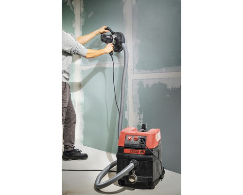 Man schuurt een wand met een gipsplaatschuurmachine die is aangesloten op een stofzuiger.