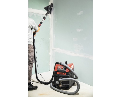 Een persoon schuurt een muur met een gipsplaatschuurmachine en een stofzuiger.