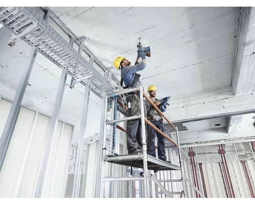 Twee bouwvakkers op een steiger installeren een kabelgoot aan het plafond van een casco gebouw.