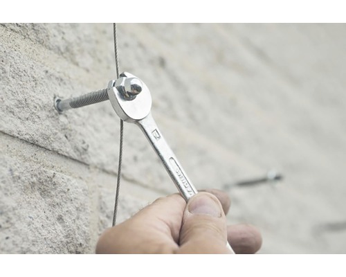 Iemand bevestigt met een moersleutel een stalen kabel aan een betonnen muur