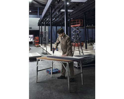 Een man boort met een accuschroevendraaier een gat in een werkstuk op een werktafel in een fabriekshal.