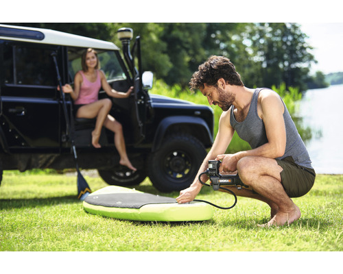 Man pompt stand-up paddleboard op met een elektrische pomp, op de achtergrond een vrouw en een terreinwagen.