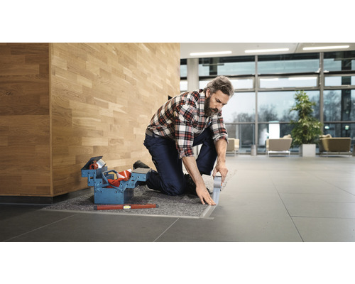 Ambachtsman knielt en verlijmt vloerrail met tape, gereedschapskist en waterpas ernaast.
