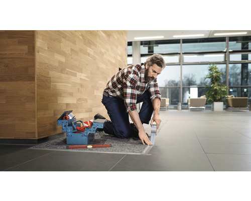 Een vakman knielt en verlijmt een vloerrail in een gebouw. Gereedschapskist en waterpas liggen klaar.