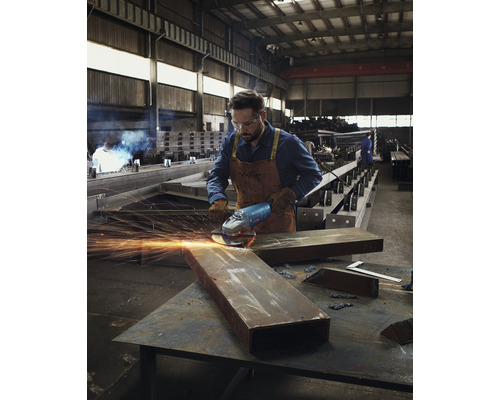 Een man in werkkleding gebruikt een haakse slijper op een stuk metaal in een werkplaats.
