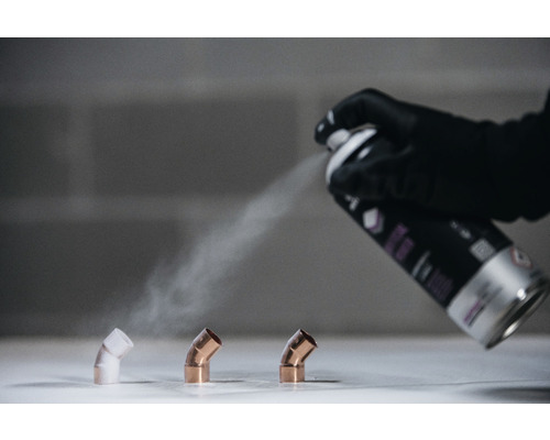 Een hand met een handschoen spuit verf op koperen bochtstukken met een spuitbus.