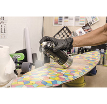 Een persoon draagt handschoenen en spuit acrylvernis op een skateboard.