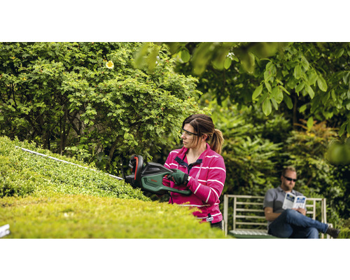 Vrouw snoeit heg met heggenschaar in de tuin, terwijl een man op een bank zit