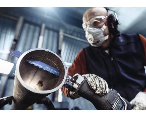 Man met veiligheidsbril, adembescherming, gehoorbescherming en handschoenen zaagt een pijp met een reciprozaag.