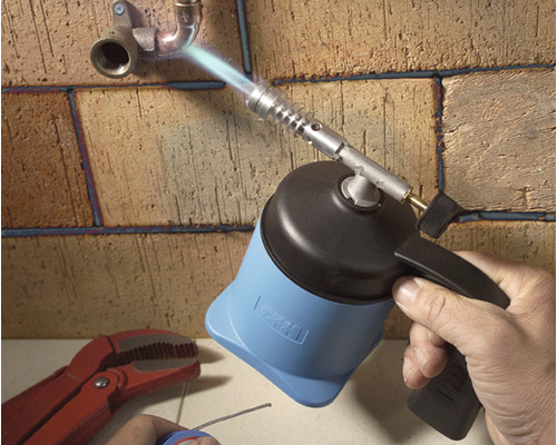 Een hand gebruikt een brander om buizen te solderen aan een bakstenen muur.