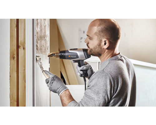 Een man verwijdert oude verf met een heteluchtpistool en plamuurmes van een raamkozijn