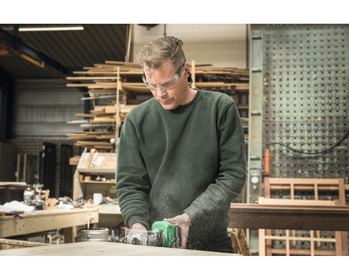 Een man werkt met een bovenfreesmachine aan een stuk hout in een werkplaats en draagt een veiligheidsbril.