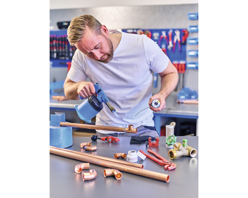 Een man soldeert koperen buizen met een brander in een werkplaats. Op de tafel liggen koperen buizen, fittingen en gereedschap.