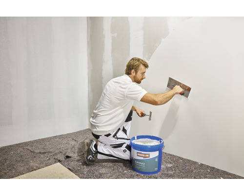 Een man brengt gladde pleister aan op een muur met een spaan. Er staat een emmer gladde pleister naast.