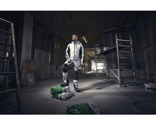 Een vakman in werkkleding staat in een casco gebouw op zakken met pleistermortel.