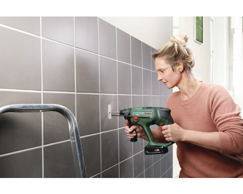 Een vrouw boort met een boormachine een gat in een betegelde muur. Ze draagt een veiligheidsbril en oordopjes.