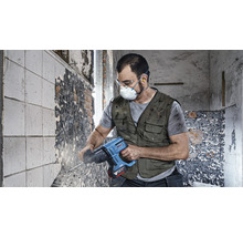 Een arbeider met veiligheidsbril, masker en gehoorbescherming gebruikt een boorhamer op een muur.