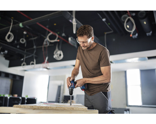 Een man boort met een boormachine een gat in een houten plank, terwijl hij een veiligheidsbril draagt.