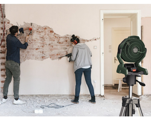 Bouwplaats met twee personen aan het renoveren, op de voorgrond een ventilator op een statief