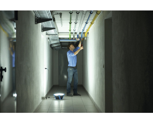 Een man werkt in een gang met een draagbare bouwlamp aan het repareren van leidingen aan het plafond