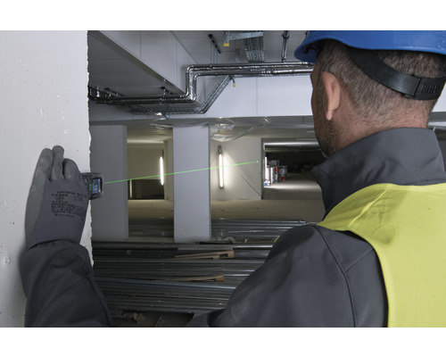 Een man met een veiligheidshelm en handschoenen gebruikt een laser afstandsmeter op een bouwplaats om een groene laserlijn op een muur te projecteren.