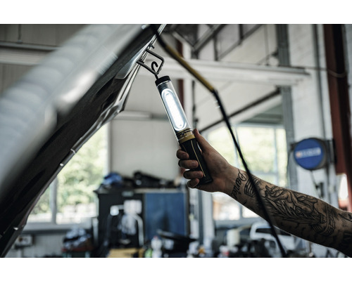 Werkplaats scène met een hand die een staaflamp onder een open motorkap houdt.