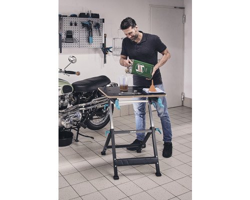 Man giet olie over op werkbank in garage, op de achtergrond een motorfiets en gereedschapswand.