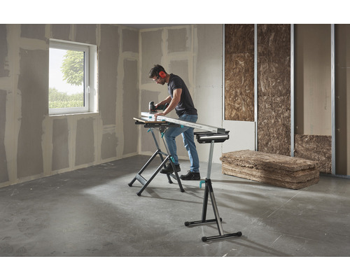 Een man werkt met een decoupeerzaag aan een werkstuk op een Wolfcraft werkbank in een ruimte met gipsplaten en isolatiemateriaal.