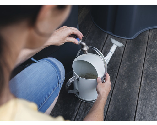 Een persoon vult een witte gieter bij de kraan in een tuin.