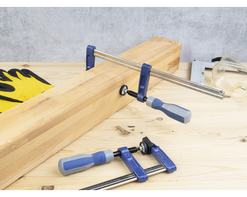 Twee lijmklemmen fixeren een stuk hout op een werkbank, aangevuld met werkhandschoenen, een tacker en een veiligheidsbril.