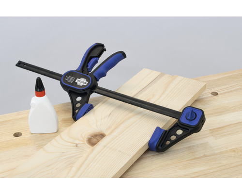 Een INDUSTRIAL éénhandklem fixeert een houten plank op een werkbank, naast een fles houtlijm.