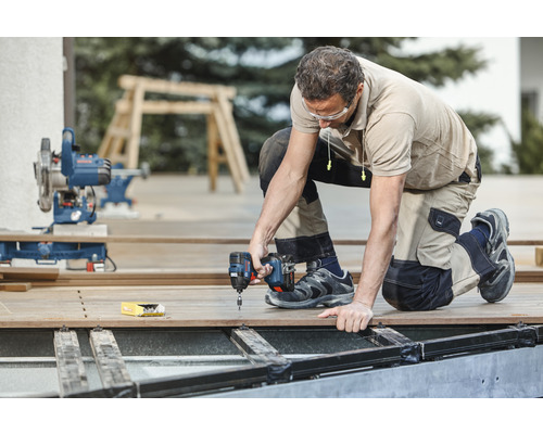Man werkt met een accuschroevendraaier aan een houten terras in de tuin. Op de achtergrond zijn een afkortzaag en een schraag te zien.