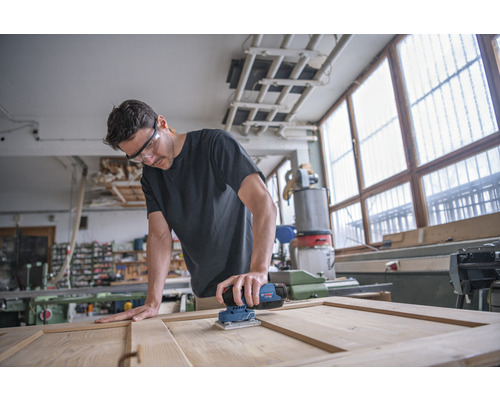Man schuurt houten deur met een elektrische schuurmachine in een werkplaats.