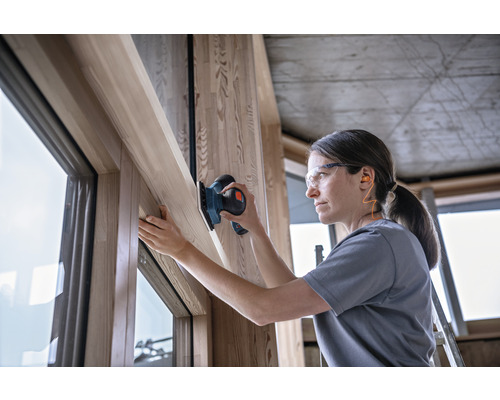 Vrouw schuurt houten raamkozijnen met een schuurmachine, terwijl ze een veiligheidsbril en oordopjes draagt.